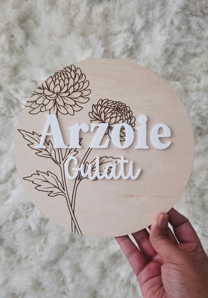 A hand holds a round wooden plaque featuring laser-engraved floral designs and the personalized name  in raised white lettering
