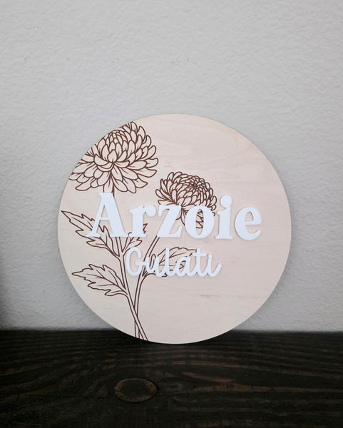 A round wooden sign with laser-engraved floral designs and the personalized name in white, displayed on a dark wooden surface against a light gray wall.